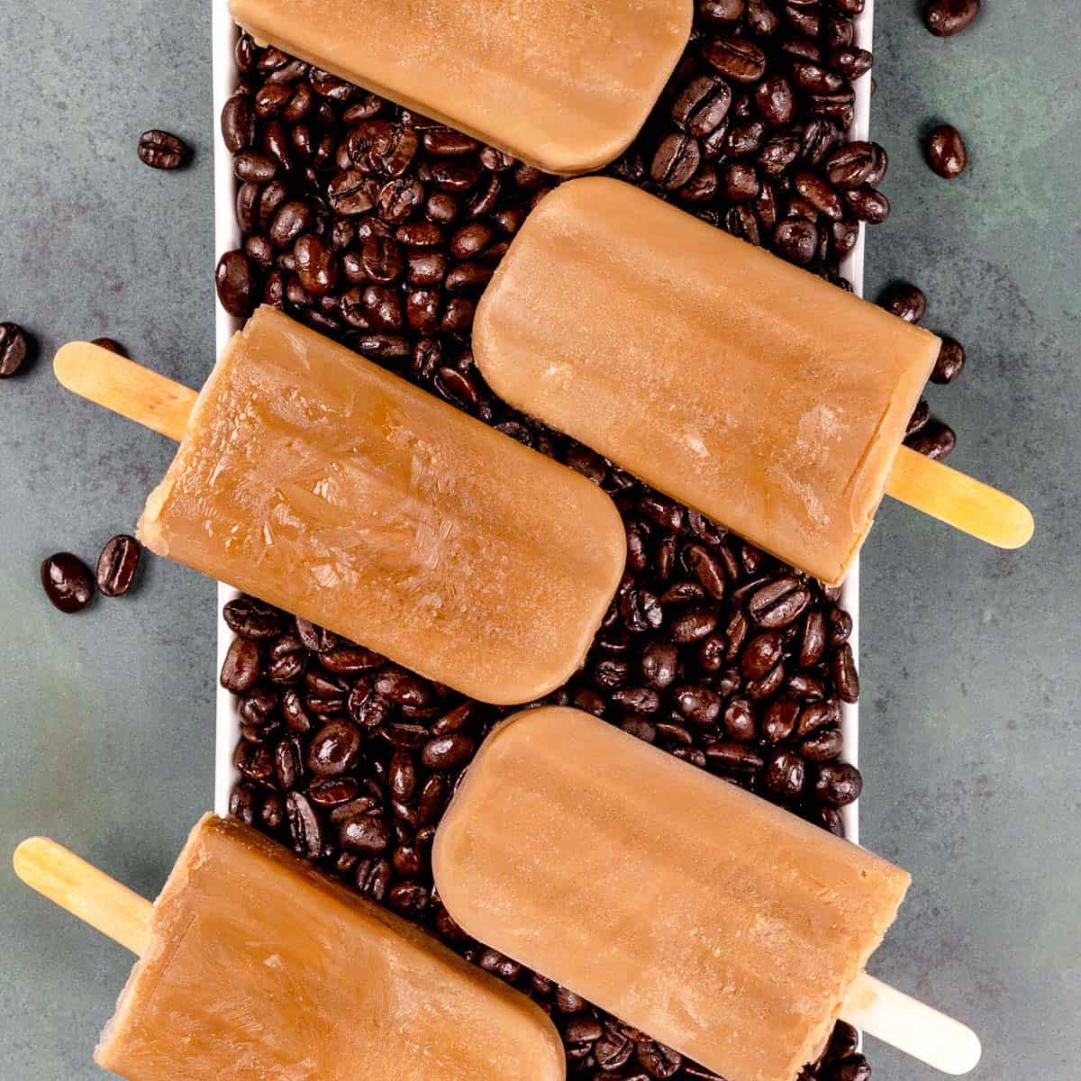 Closeup of 5 coffee popsicles on a long white serving tray filled with roasted coffee beans.
