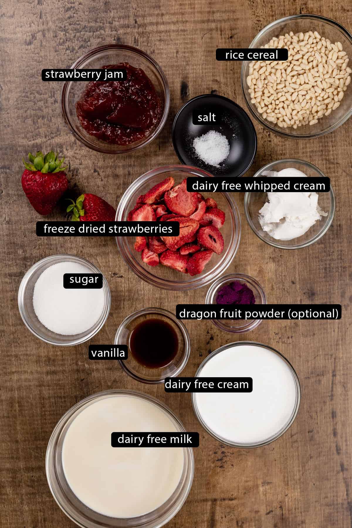 Ingredients for strawberry shortcake ice cream bars in various glass bowls on a wood table. Black and white labels have been added to name each ingredient like strawberries and vanilla.