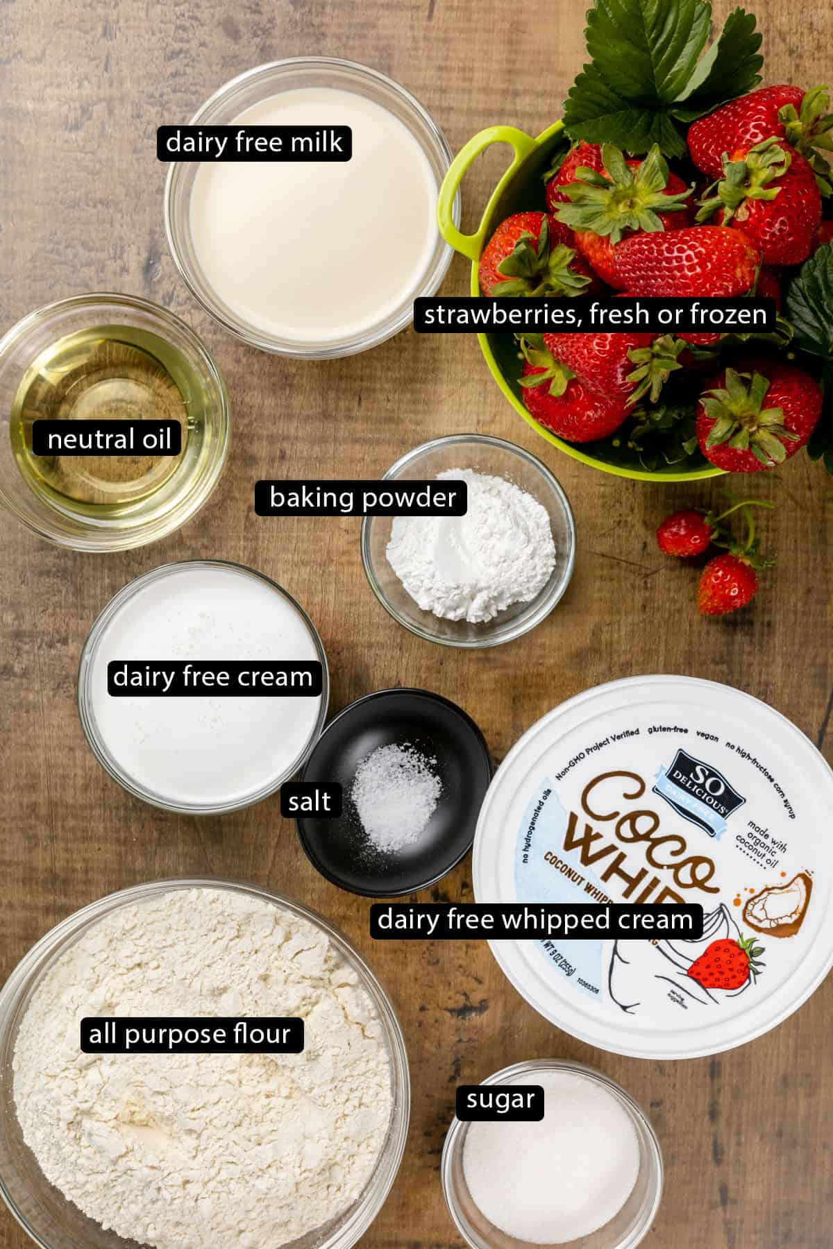 Ingredients for vegan strawberry shortcakes in various glass bowls on a wood tabletop. Black and white labels have been added to name each ingredient like flour, sugar, and baking powder.