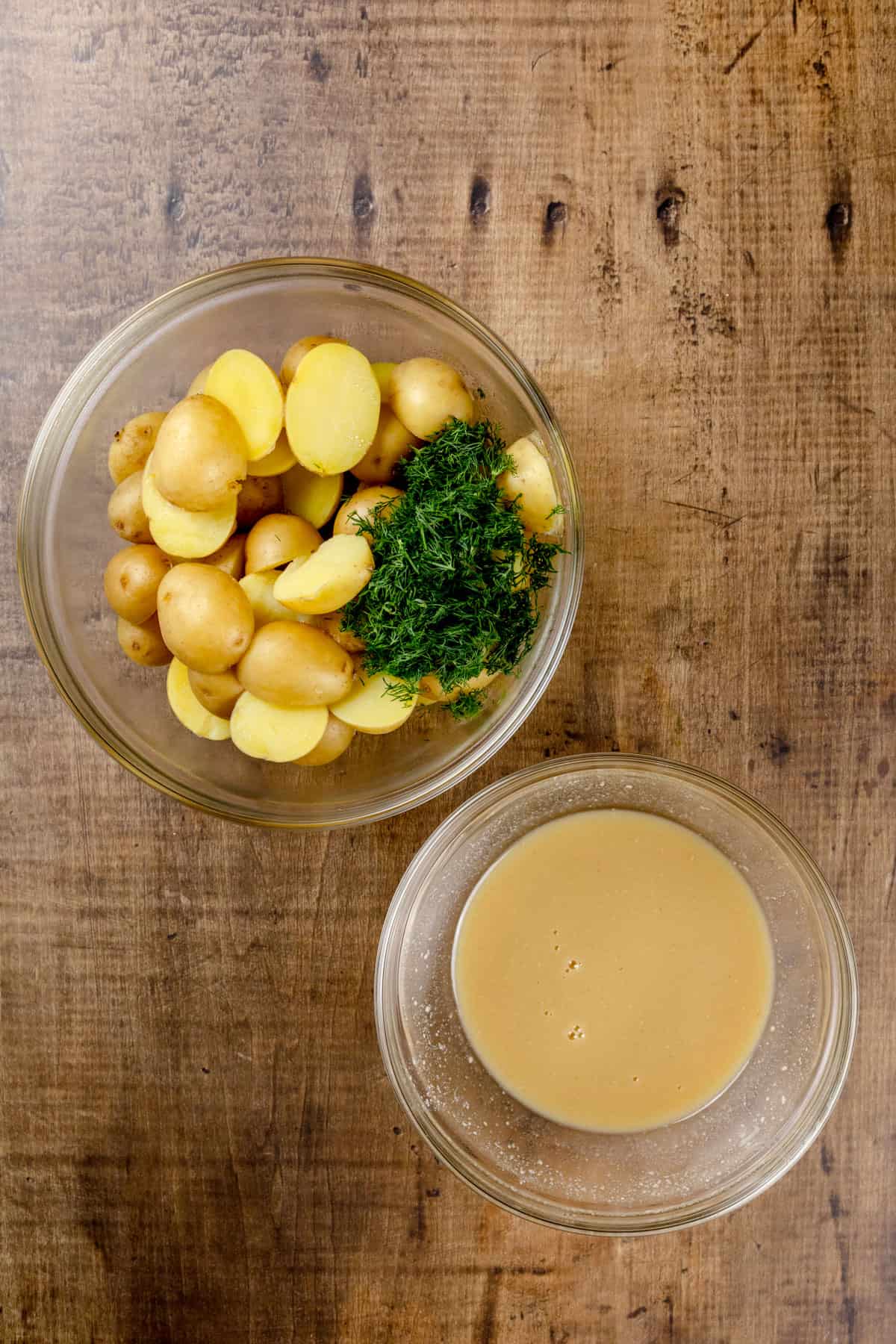Two glass bowls on a wood tabletop filled with the dressing in one and the cooked potatoes in the other.