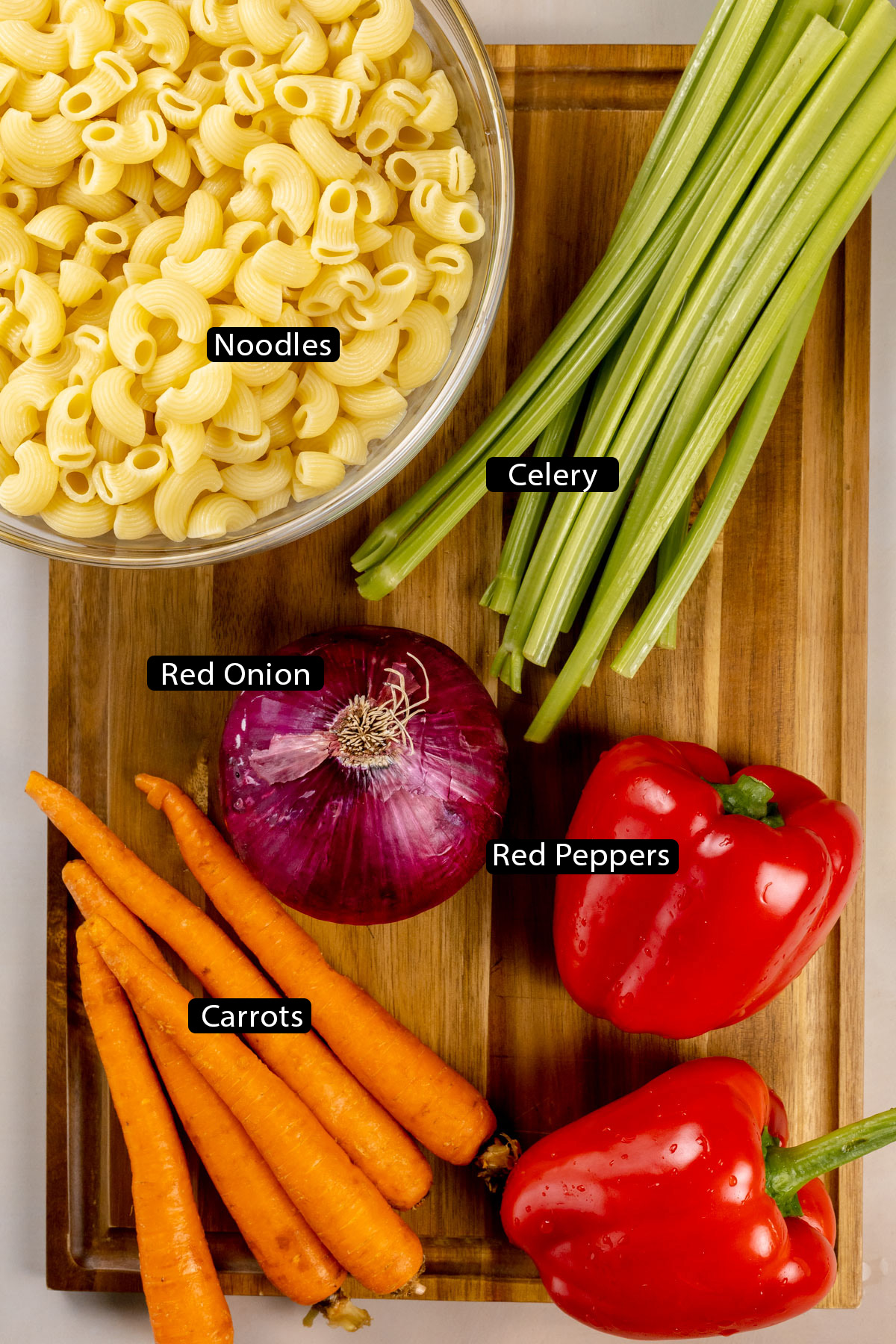 Ingredients for the vegan macaroni salad on a wood cutting board. Black and white labels have been added to name each ingredient like noodles and red peppers.
