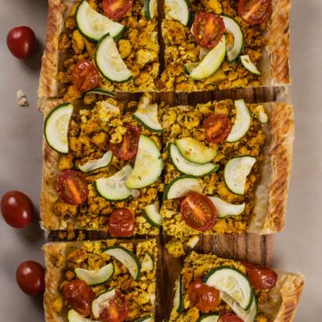 Close up of 6 slices of a tofu scramble tart on a wood cutting board. The tofu, zucchini, and tomatoes are all crispy.