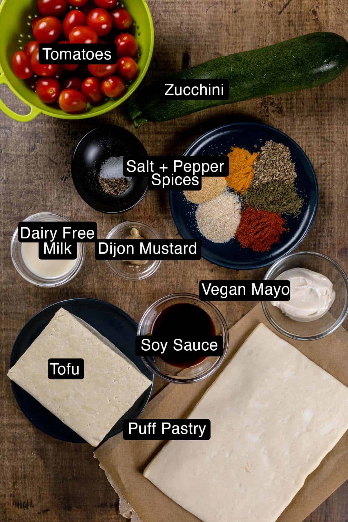 Ingredients for the tofu scramble tart in various glass bowls on a wood table top. Black and white labels have been added to name each ingredient.