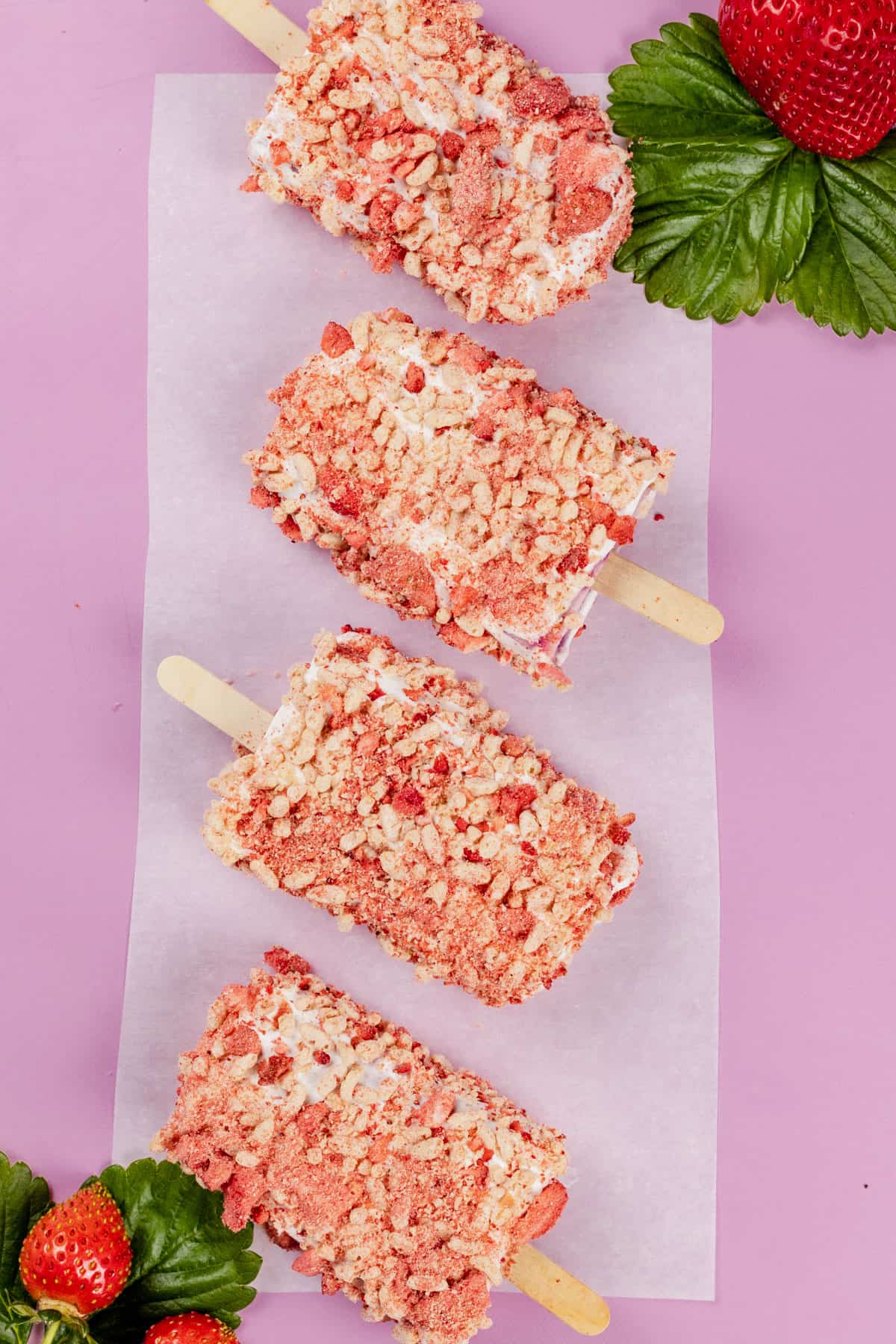Many dairy free strawberry shortcake ice cream bars on a bright pink background with a few fresh berries beside them.