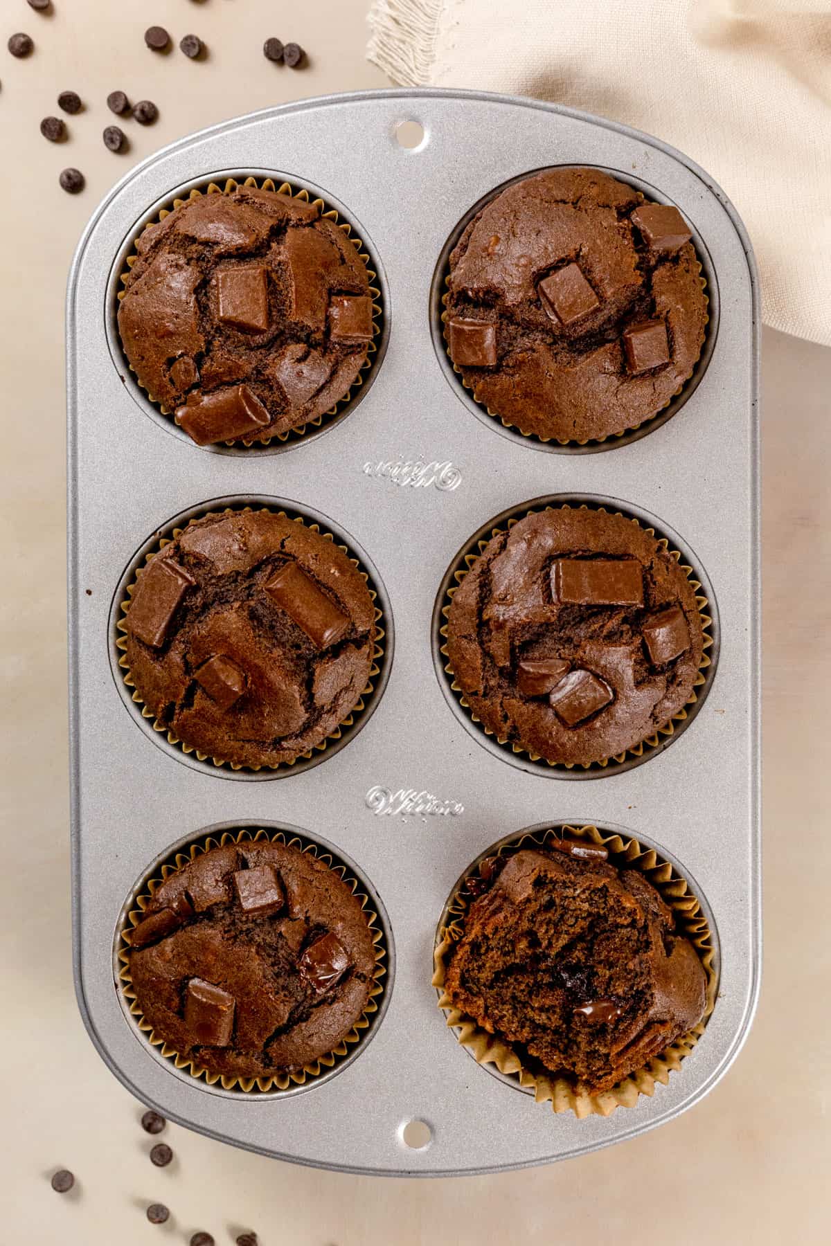 Chocolate muffins in a silver muffin tray after baking. One has been cut in half to show the fluffy texture inside. A few chocolate chips are next to the tray.