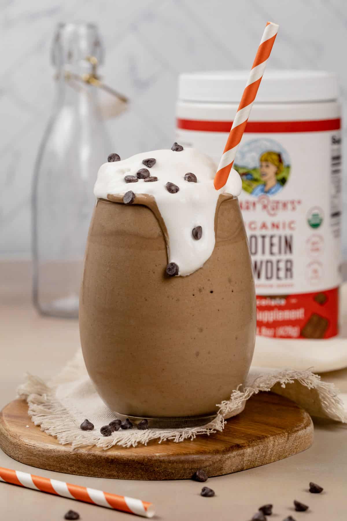 A glass filled with the chocolate protein shake and with a dollop of whipped cream on top that is artfully overflowing in the glass. Tiny chocolate chips are sprinkled on top. A white and orange stripe straw is in the glass.