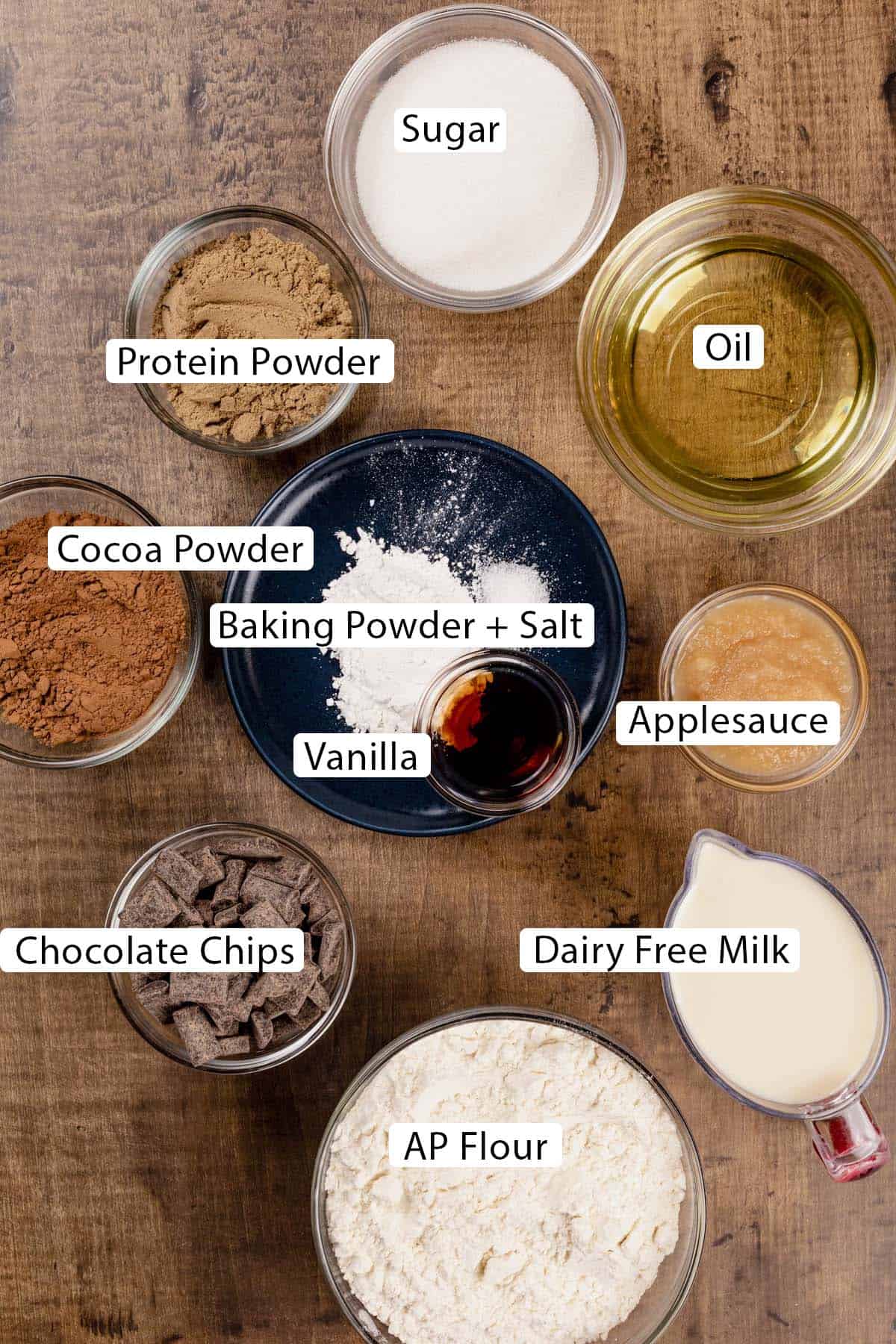 Ingredients for chocolate muffins in various glass bowls on a wood tabletop. Black and white labels have been added to name each ingredient.