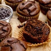 Chocolate protein muffins on a table with a small bowl of chocolate chips. One muffin up front has been opened so you can see the rich chocolatey inside.