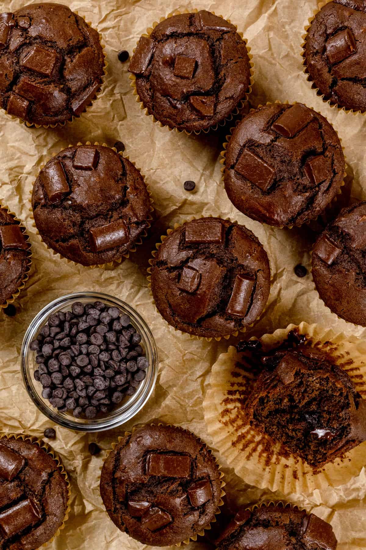 Overview of chocolate muffins on a wrinkled parchment paper. A bowl of mini chocolate chips is next to the muffins.