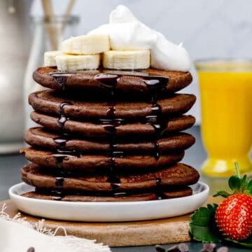 A stack of chocolate protein pancakes on a white plate topped with banana slices, chocolate sauce, and whipped coconut cream. Fresh berries and juice are blurred in the background.