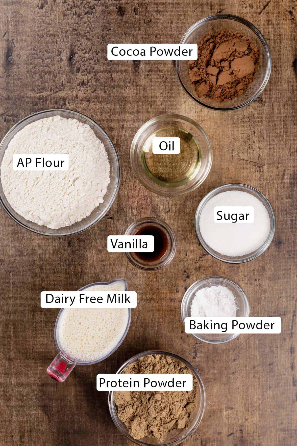 Ingredients for chocolate protein pancakes in several glass bowls on a wood tabletop. Black and white labels have been added to name each ingredient.