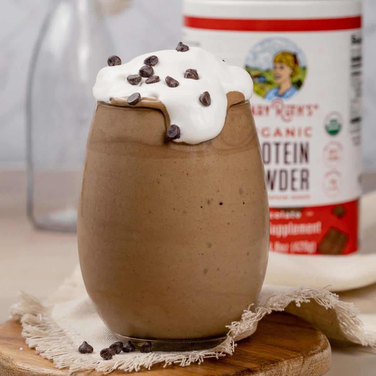 A glass filled with chocolate protein shake with whipped cream and mini chocolate chips on top. More chips and a few straws are next to the glass. The bottle of protein powder is blurred in the background.