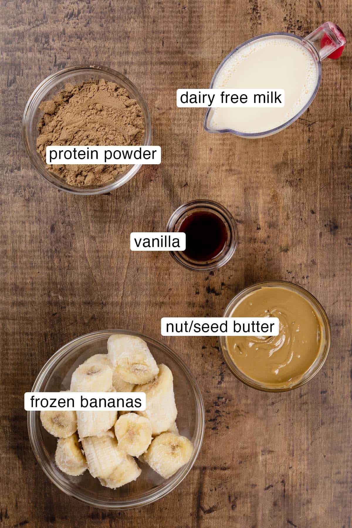 Ingredients for a nut butter chocolate banana protein shake in various glass bowls on a kitchen tabletop. Black and white labels have been added to name each ingredient.