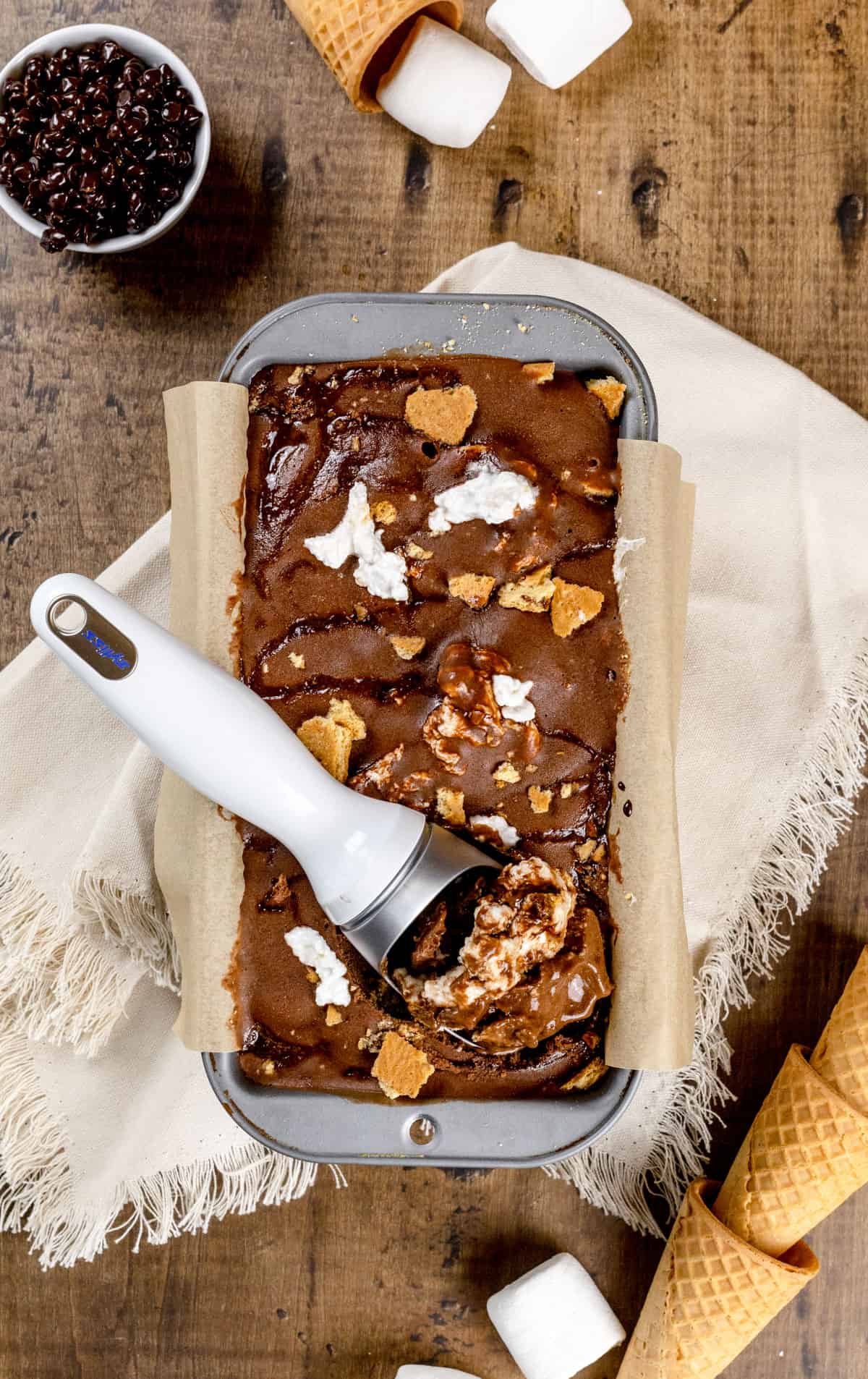 A metal loaf pan filled with the finished and fully frozen s'mores nice cream. Ice cream cones, marshmallows, and chocolate chips are next to the pan.