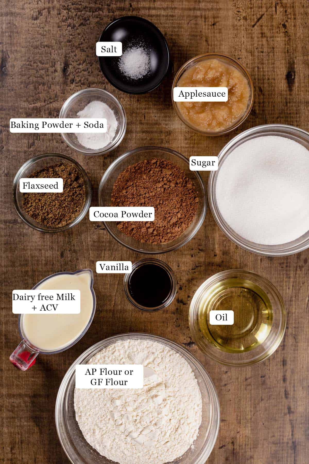 Ingredients for a vegan chocolate sheet cake in various glass bowls on a wood table. Black and white labels have been added to name each ingredient like flour, cocoa powder, and applesauce.