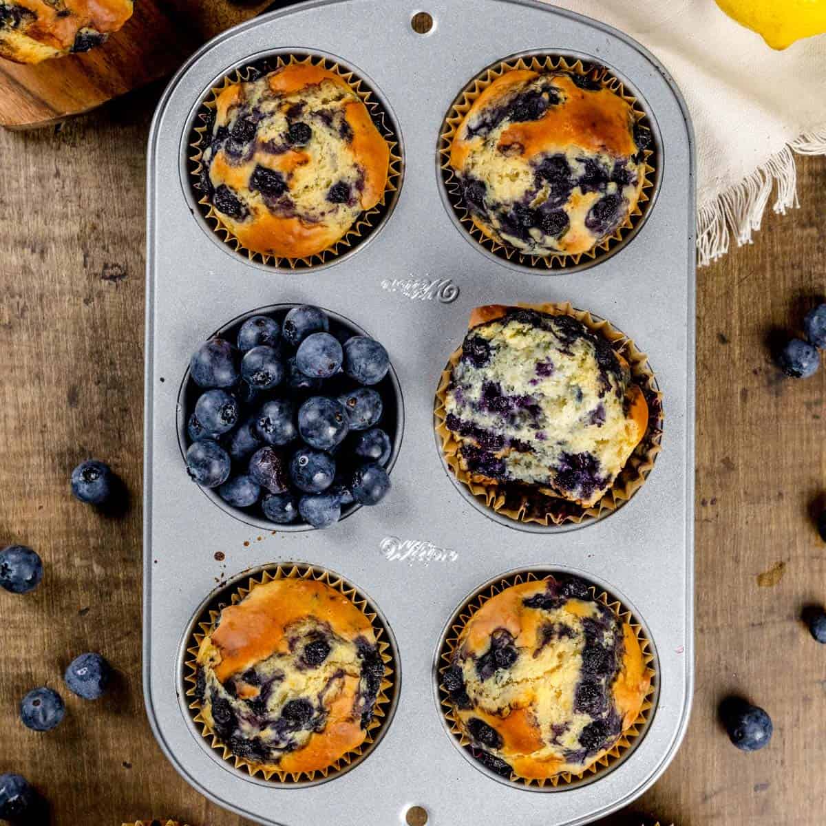 Protein blueberry muffins in a silver muffin tray on a wood tabletop. More muffins and fresh blueberries are surrounding the tray.