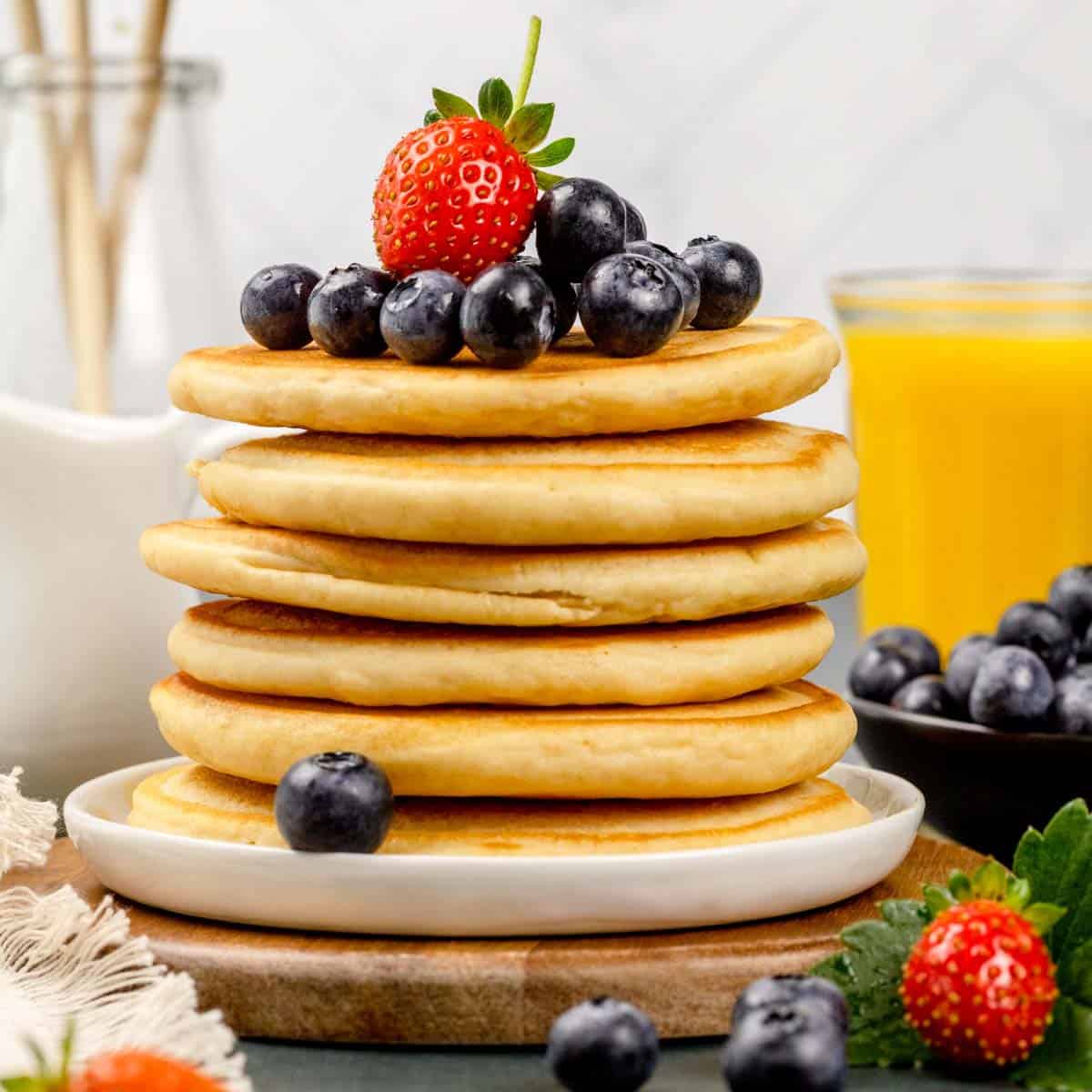 Tall stack of protein pancakes topped with fresh berries on a white plate. More fresh berries surround the plate and a glass of orange juice is blurred in the background.