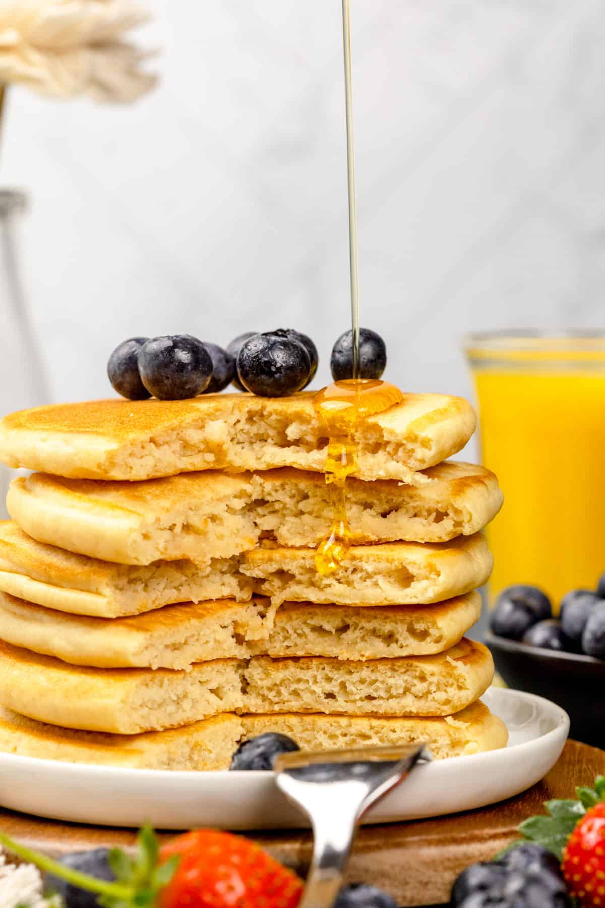 Stack of protein pancakes on a white plate with a large bite taken out of the stack. Pancake syrup is being poured on top.
