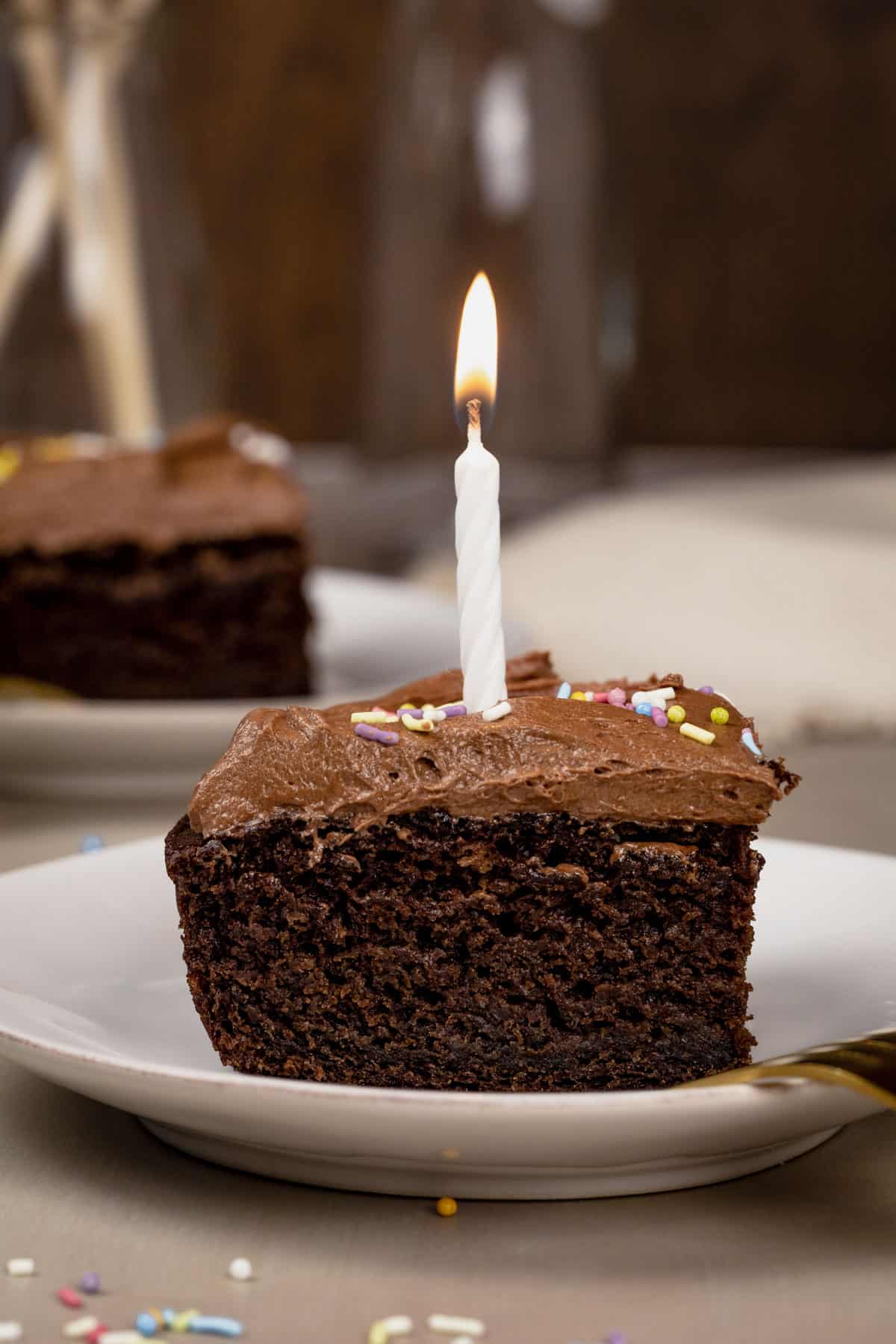 A slice of chocolate sheet cake on a small plate with a white candle on top that is lit. Sprinkles are on top of the cake and scattered around the plate. More slices are blurred in the background.