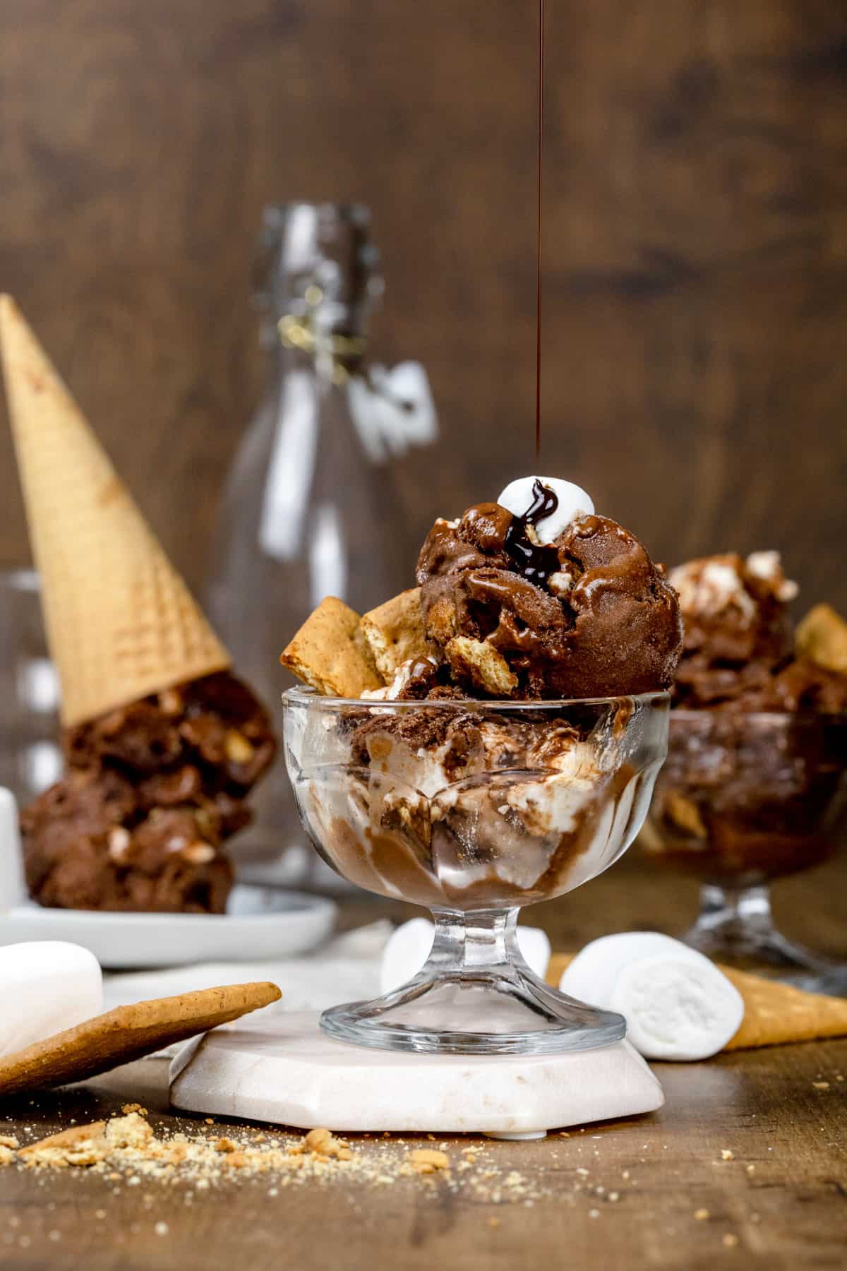 A glass bowl filled with s'mores nice cream with a drizzle of chocolate sauce being poured from above the bowl.