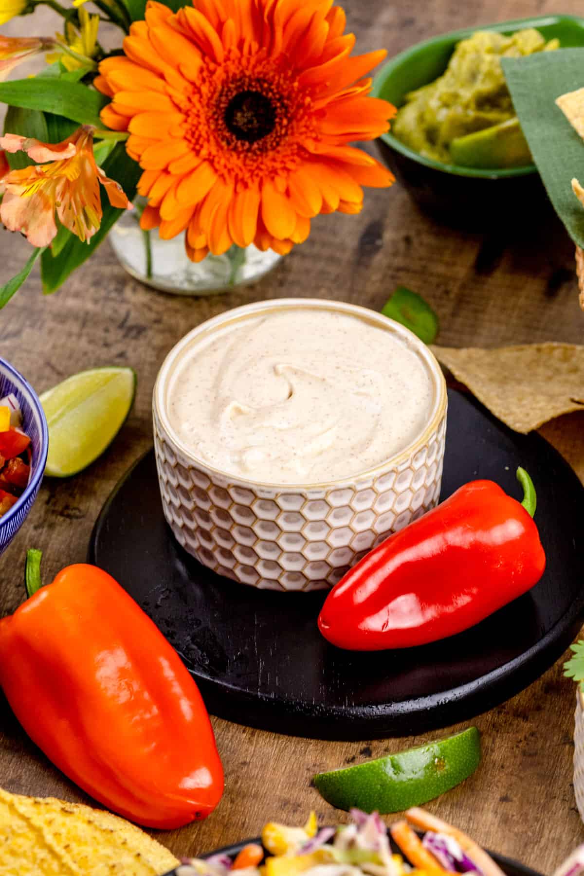 A bowl is filled with dairy free chipotle crema sauce. Peppers, lime wedges, fresh flowers, and more surround the bowl on the tabletop.