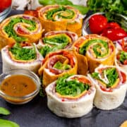 Closeup of many dairy free Italian pinwheels in tomato wraps and gluten free wraps. They are on a black slate serving tray with tiny bowls of dip and peppers.