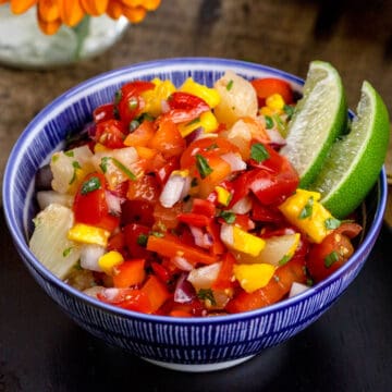 A blue bowl filled with fresh mango pineapple salsa. Two wedges of lime are in the bowl as well.