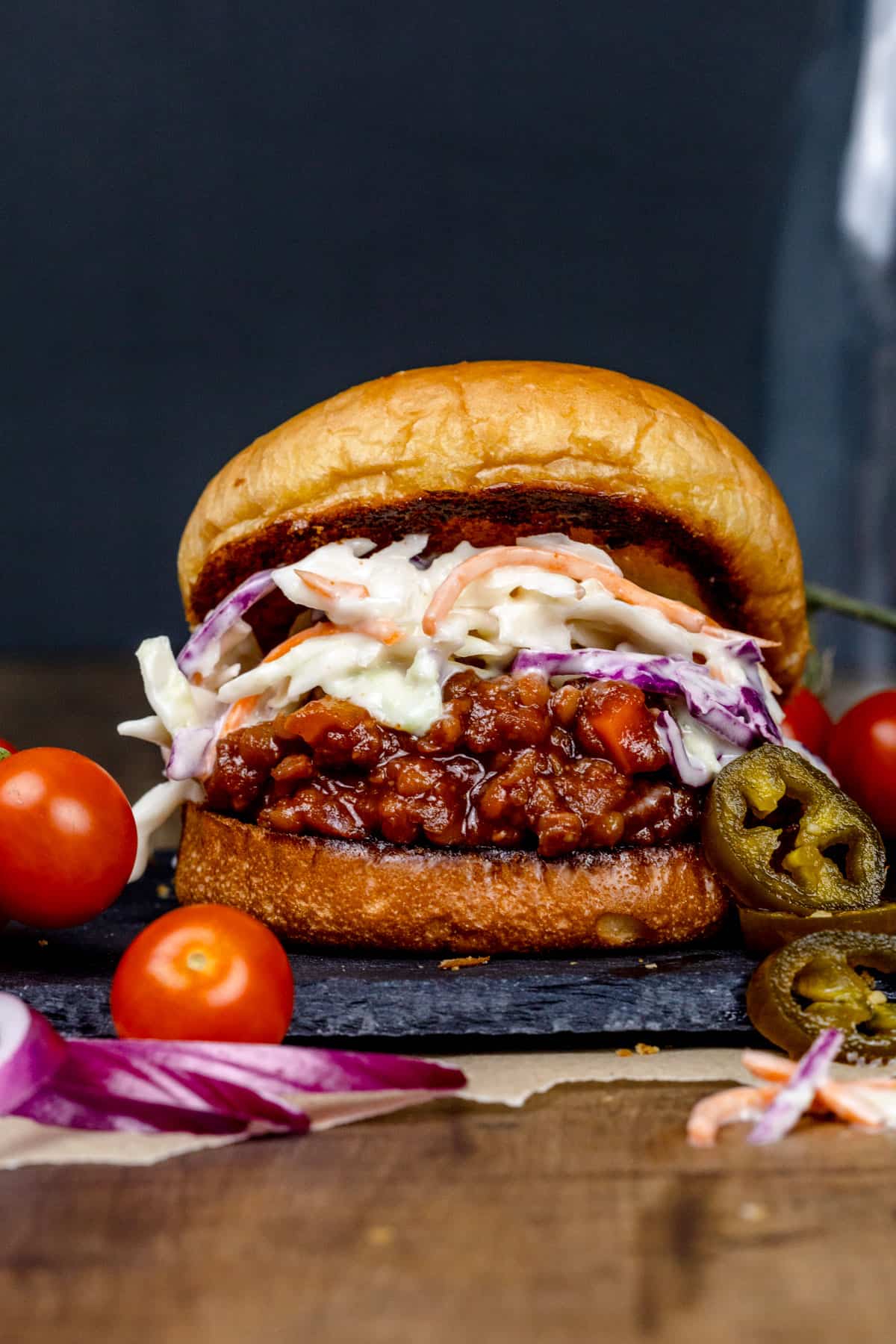 A bbq lentil burger on a dark slate serving tray. The bun is tilted back and you can see the coleslaw on top of the lentils. tiny tomatoes, pickles jalapeños, and red onion slices are next to the burger.