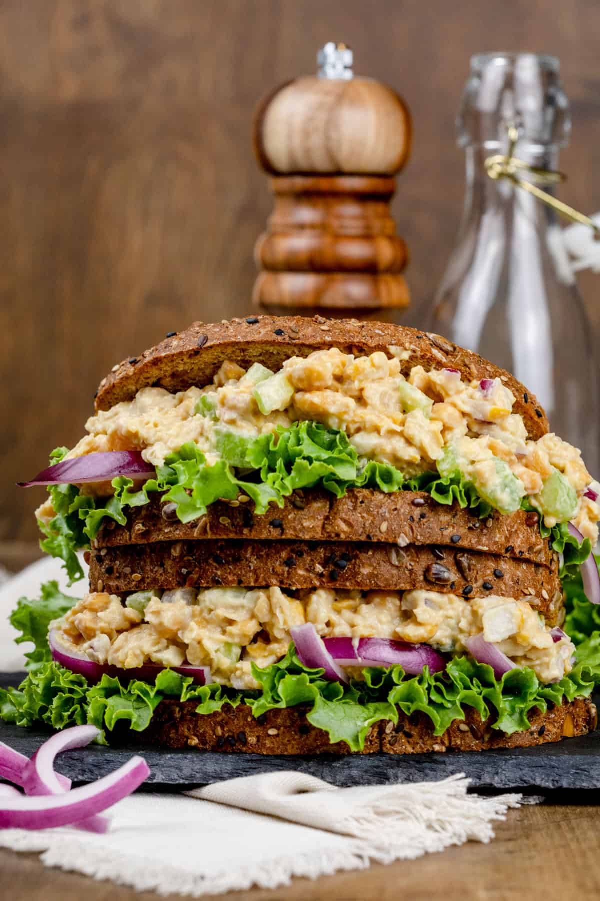 Two sandwiches stacked on top of each other filled with a mock tuna chickpea of the sea filling on hearty brown bread. Slices of red onion are next to the sandwiches. In this image, the glass jar is blurred in the background on the right side of the image.