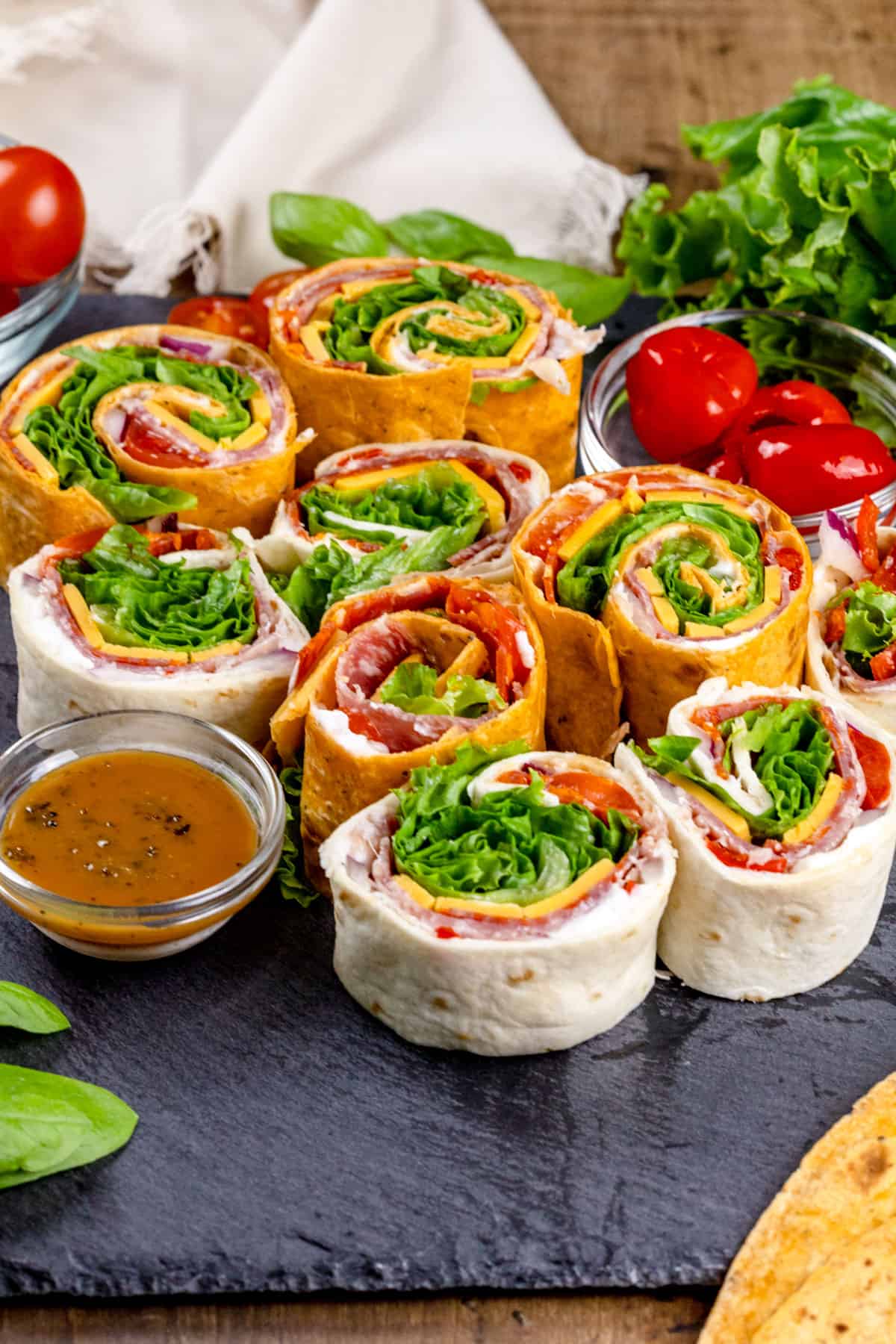 Many pinwheels on a black slate serving tray with small bowls filled with dip and more peppers. Fresh lettuce and other tomatoes are blurred in the background.