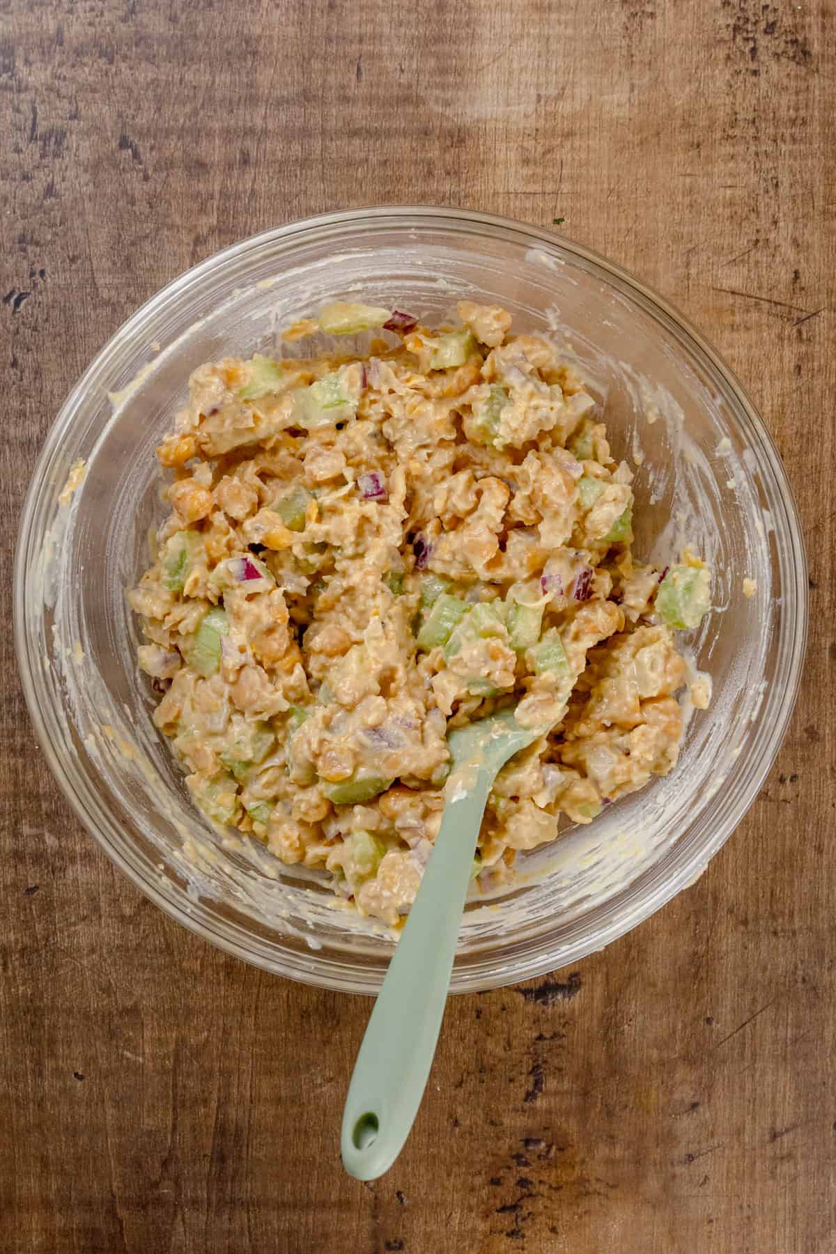 A glass bowl on a wood tabletop is filled with the finished filling for a chickpea of the sea sandwich. A green spatula is in the bowl stirring.