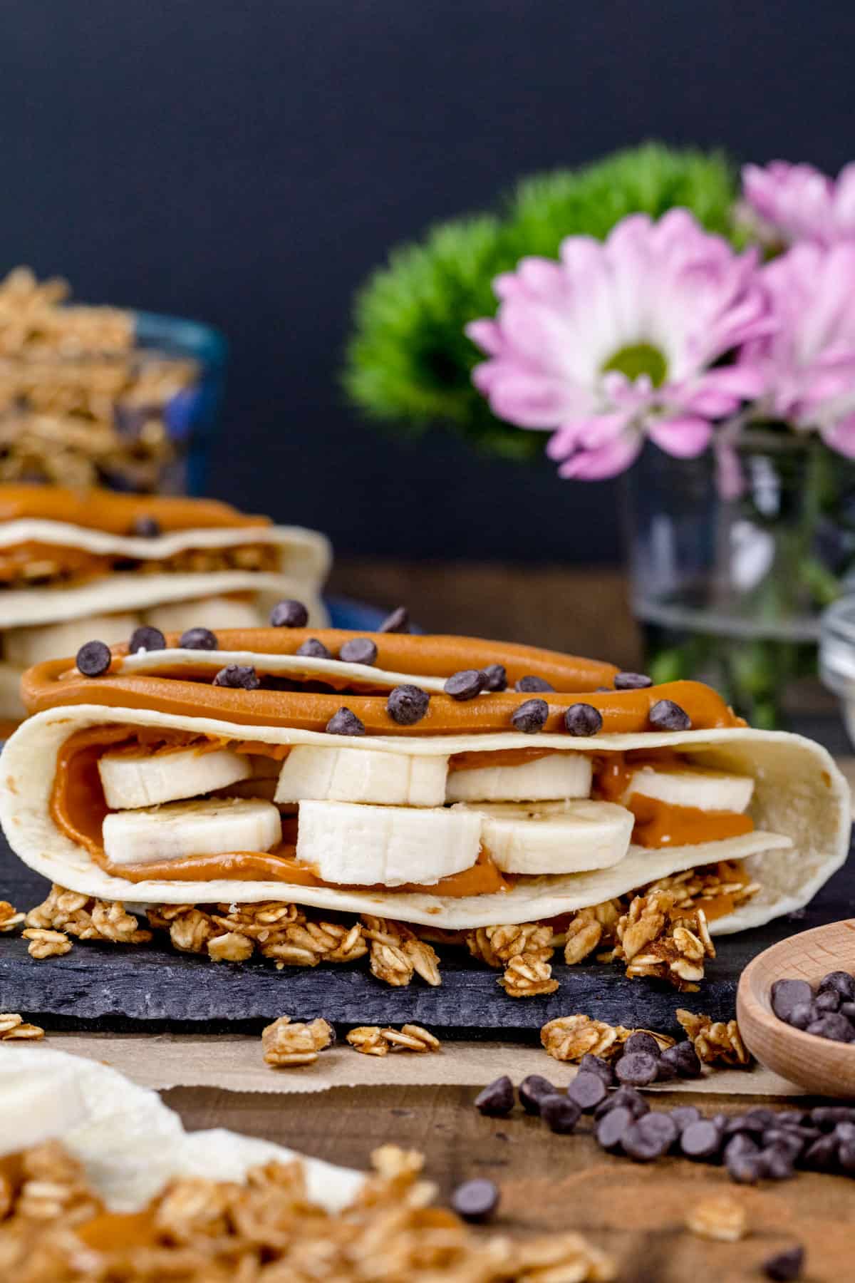 A finished sunbutter and banana wrap on a kitchen table filled with ingredients like banana slices, mini chocolate chips, and granola. Fresh flowers and more wraps are blurred in the background.