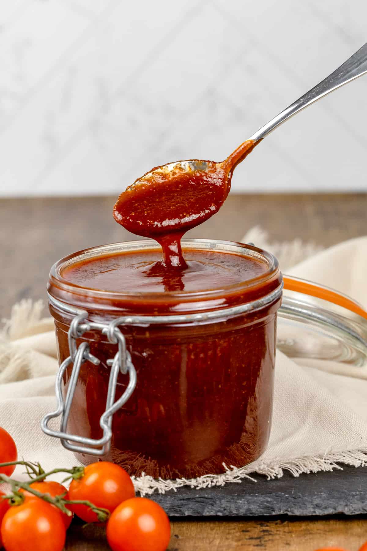 A glass jar is filled with vegan bbq sauce. A silver spoon has pulled up a spoonful of sauce and it is dripping back down into the jar. Tiny tomatoes are blurred in the foreground.