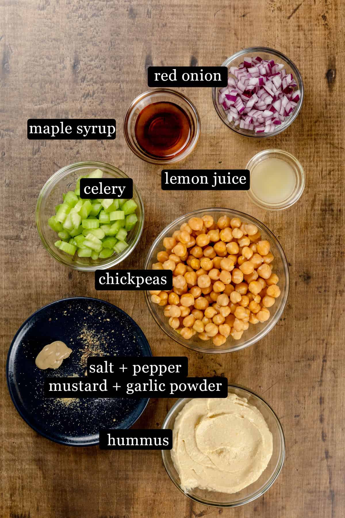 Ingredients for a chickpea of the sea sandwich in various glass bowls on a wood table. Black and white labels have been added to the image to name each ingredient, like chickpeas and lemon juice.