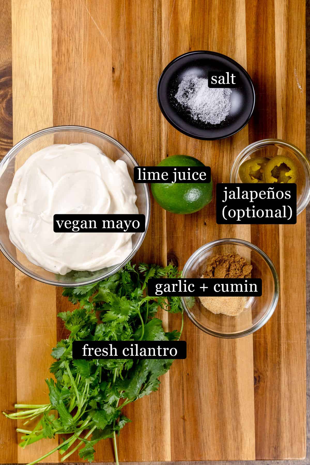 Ingredients for the cilantro aioli sauce in various glass bowls on a wood tabletop. Black and white labels have been added to name each ingredient, like fresh cilantro and vegan mayo.