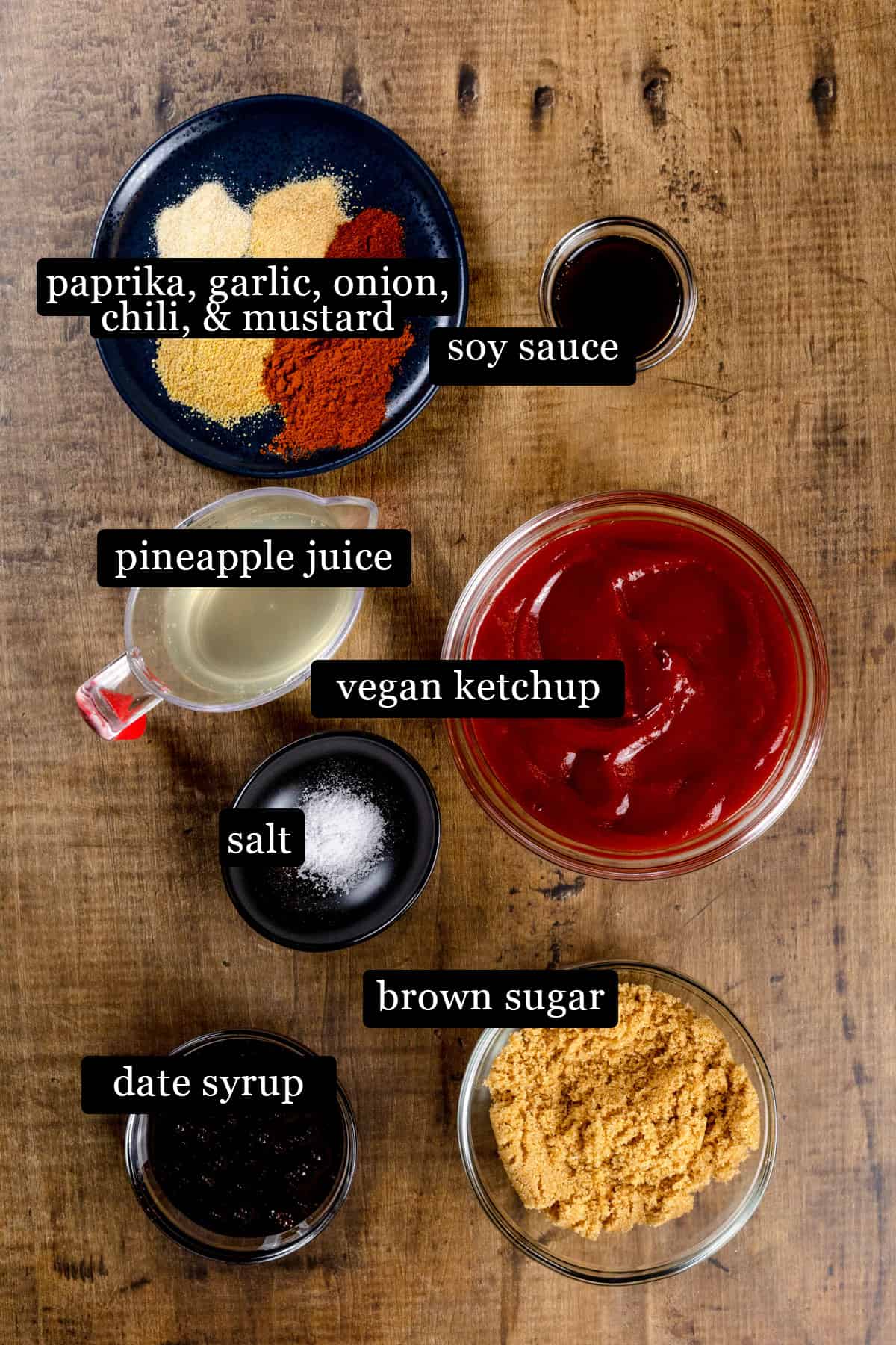 Ingredients for the bbq sauce in various glass bowls and on plates on a kitchen table. Black and white labels have been added to name each ingredient.