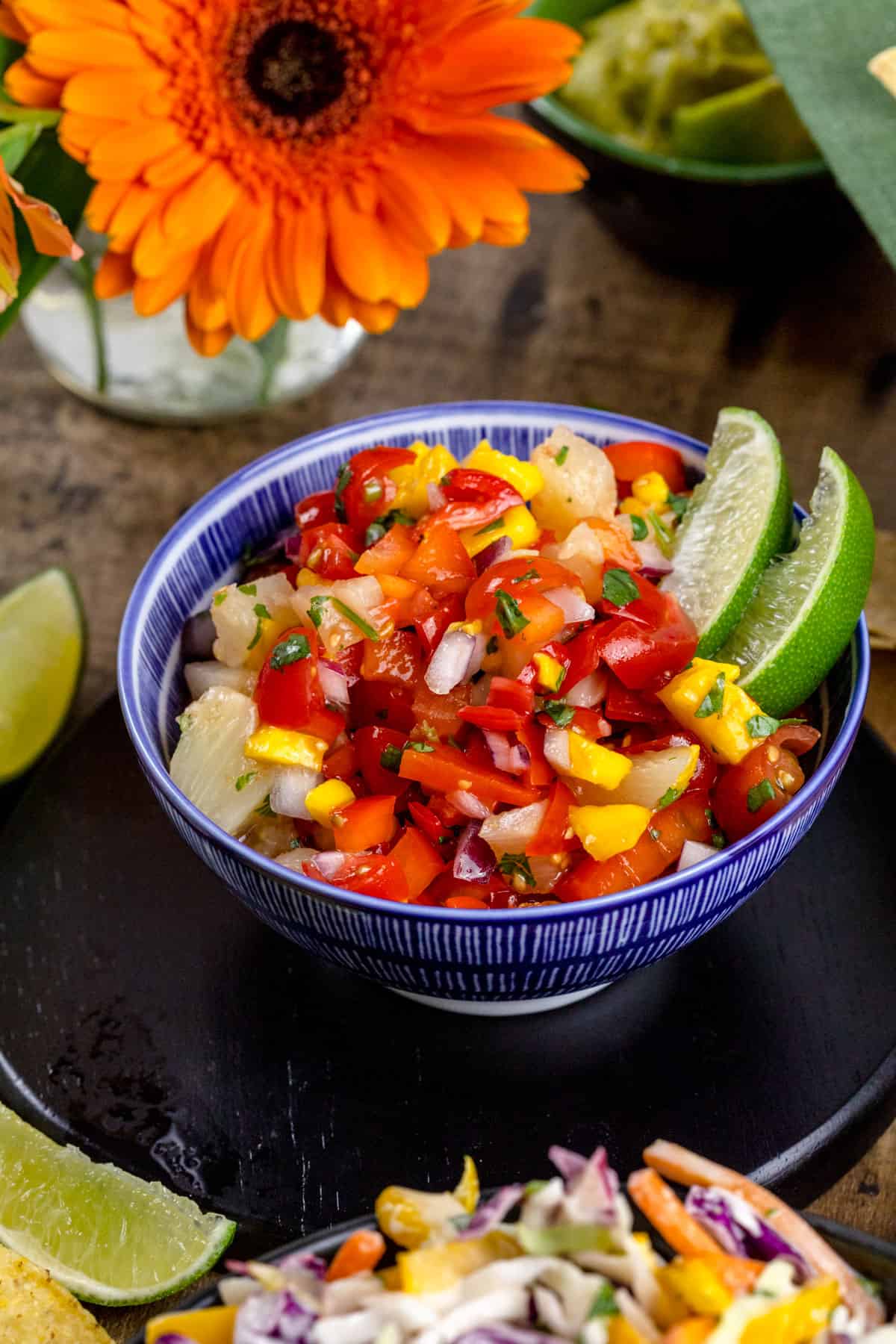 A side view of a blue bowl filled with fresh mango pineapple salsa with lime wedges and fresh cilantro. More limes, fresh flowers, and taco night ingredients surround the bowl.