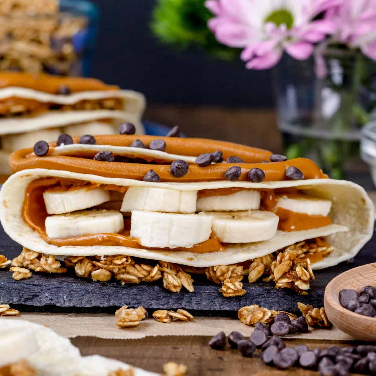 Closeup of a finished sunbutter and banana wrap on a kitchen table filled with ingredients like banana slices, mini chocolate chips, and granola. Fresh flowers and more wraps are blurred in the background.