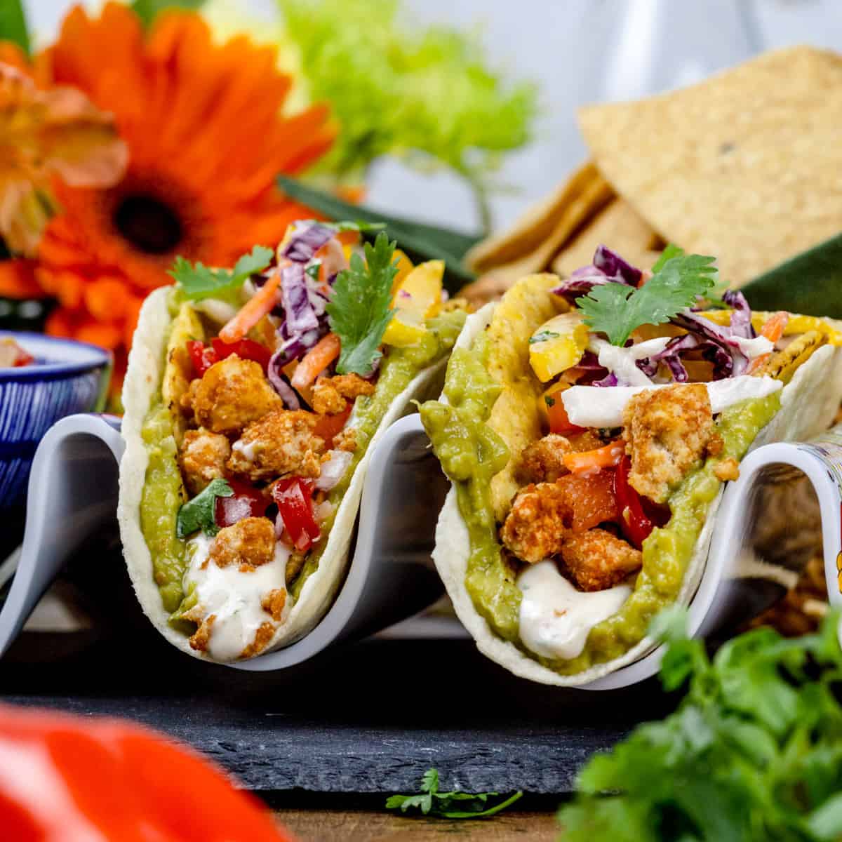 Closeup of two tofu tacos in a taco stand in both crunchy and soft shells. Toppings like guacamole, salsa, and sour cream are on top. Fresh ingredients like peppers, cilantro, and fresh flowers are surrounding the tacos.