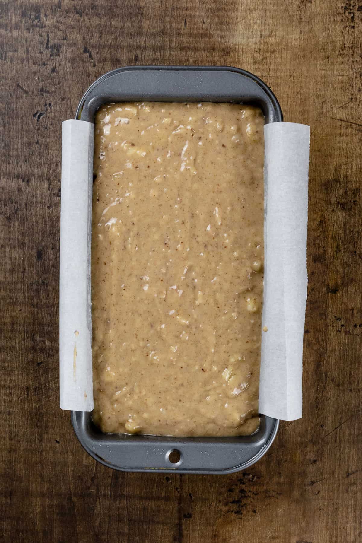 Banana bread batter is unbaked in a metal pan on a wood kitchen table.