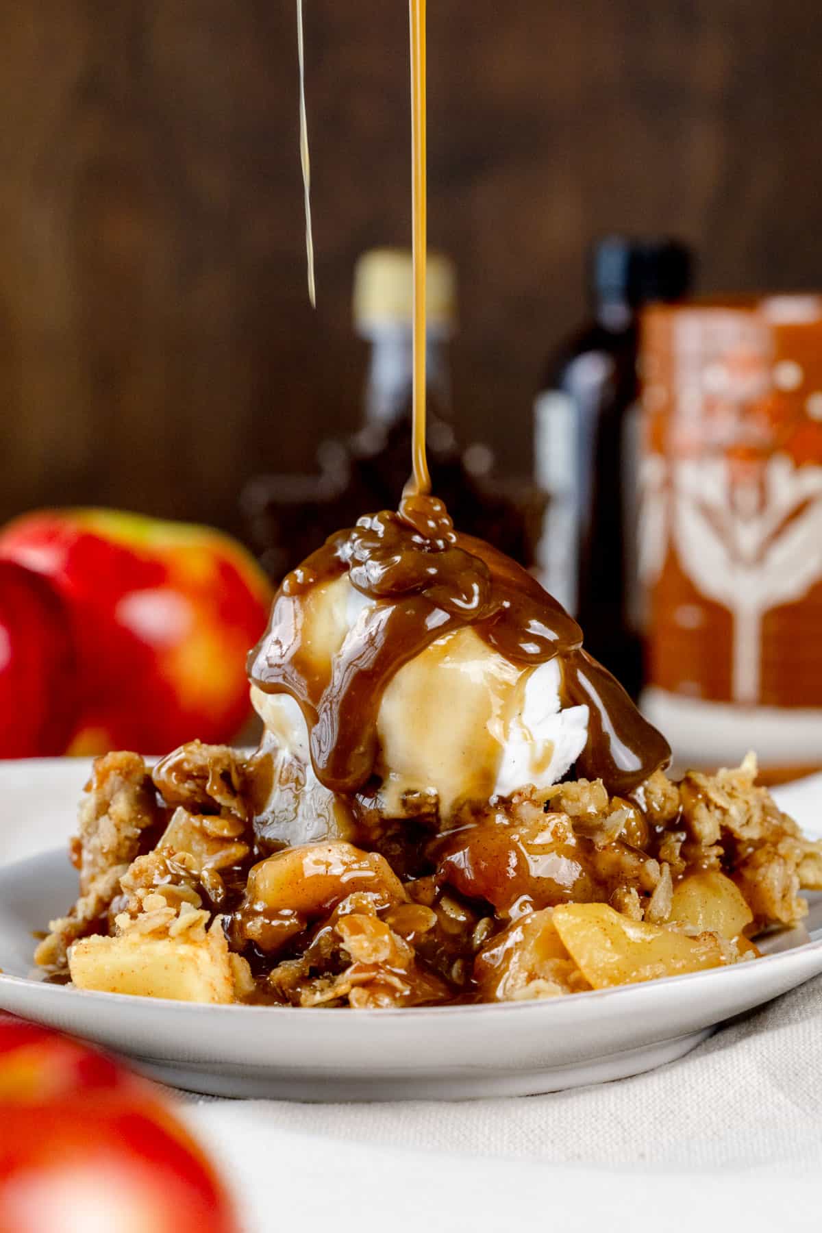 A plate covered in apple crisp with a scoop of vanilla ice cream with fresh caramel sauce being poured over top is on a kitchen table with more apples and syrup blurred in the background.