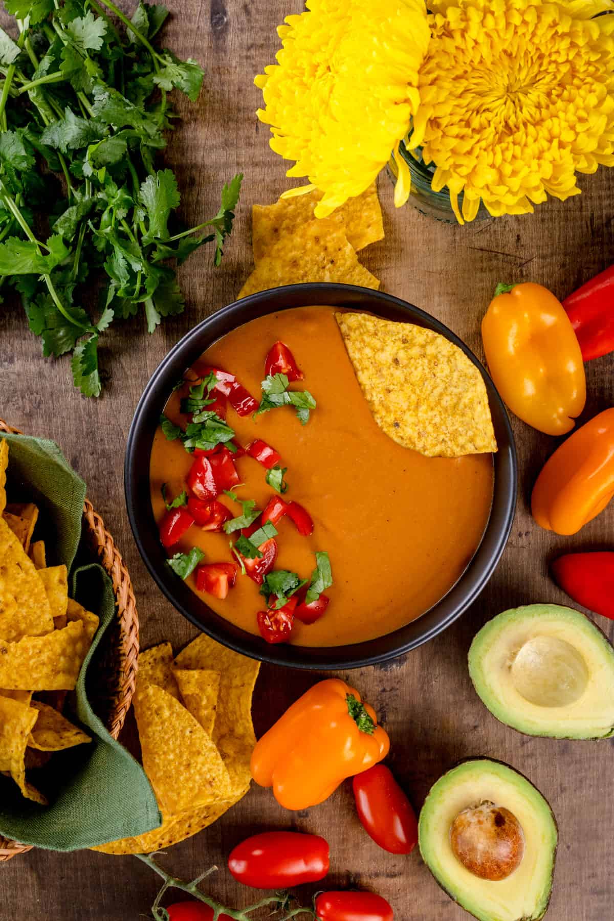 A dark bowl is filled with dairy free quest. Fresh tomato and cilantro are on top of the quest. A yellow corn chip is tucked in the side of the bowl. More chips, peppers, avocados, fresh cilantro, and fresh flowers surround the bowl.