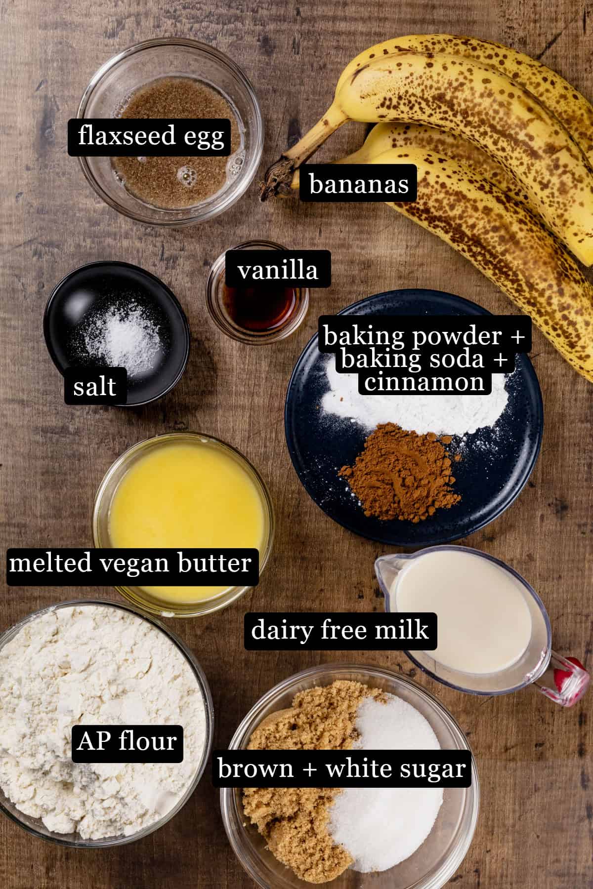 Ingredients for banana bread in various glass bowls on a wood tabletop. Black and white labels have been added to name each ingredient like bananas and dairy free milk.