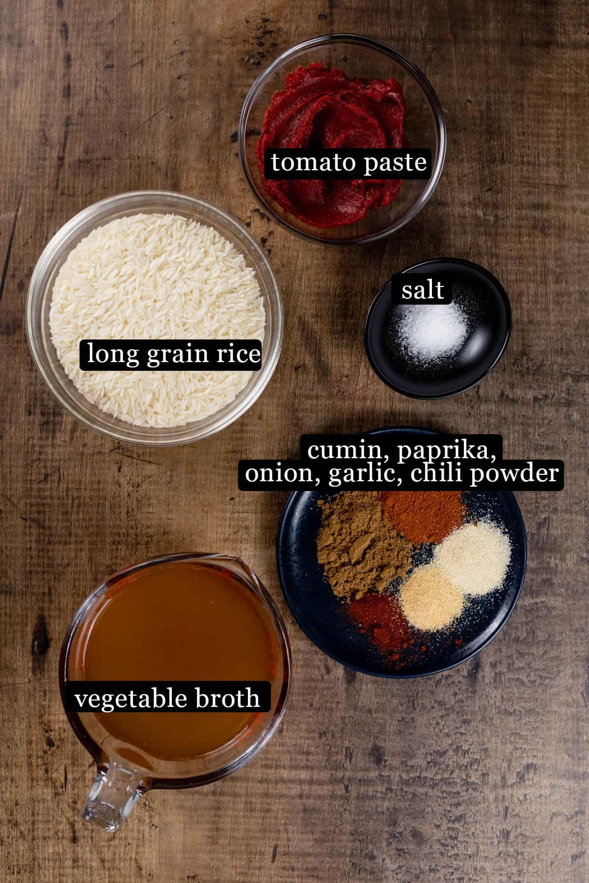 Ingredients for Spanish rice in various glass bowls on a wood tabletop. Black and white labels have been added to name each ingredient, like "tomato paste" and "long grain rice".