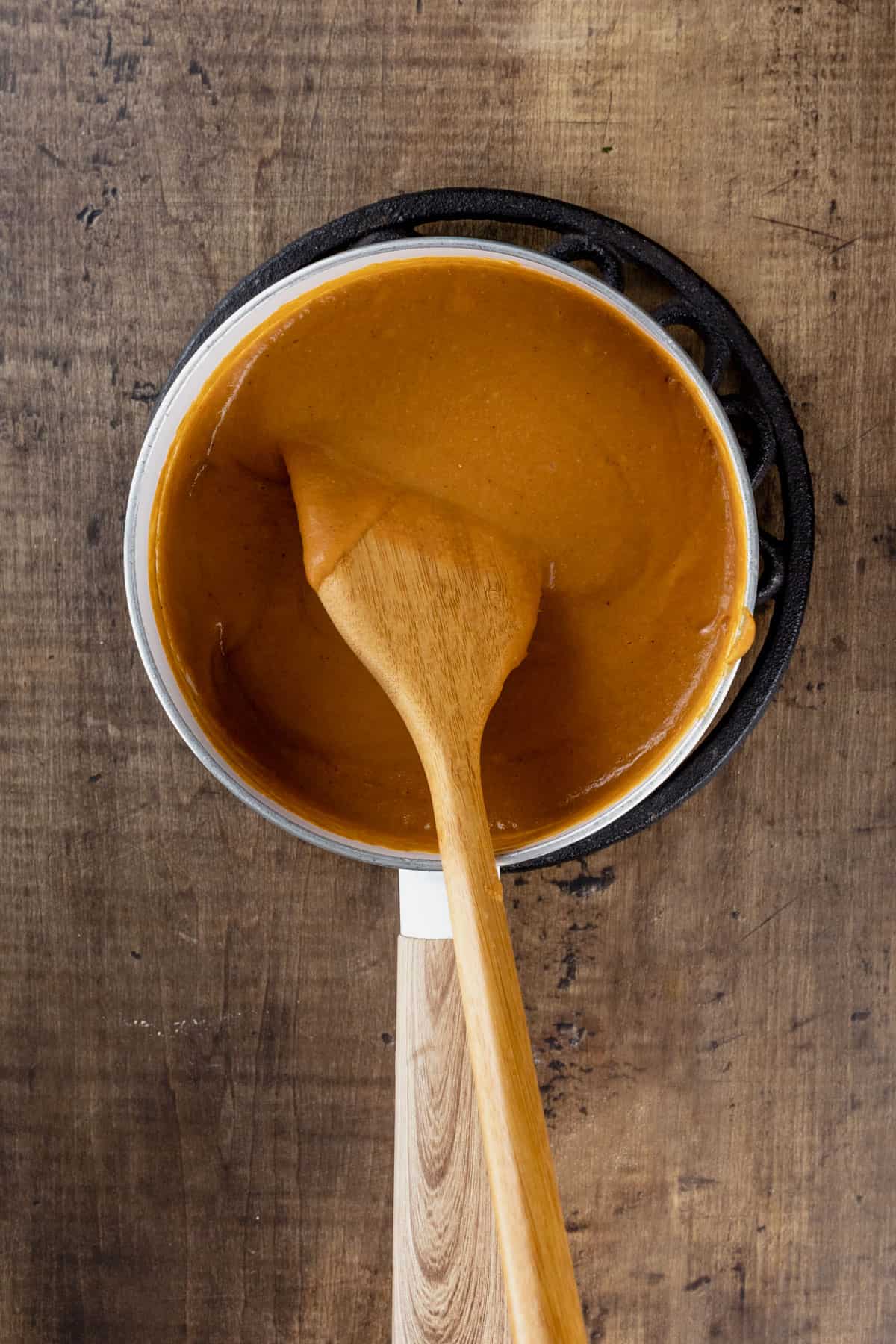 A wood spoon is stirring the thick queso sauce in a white pot on a wood kitchen table.