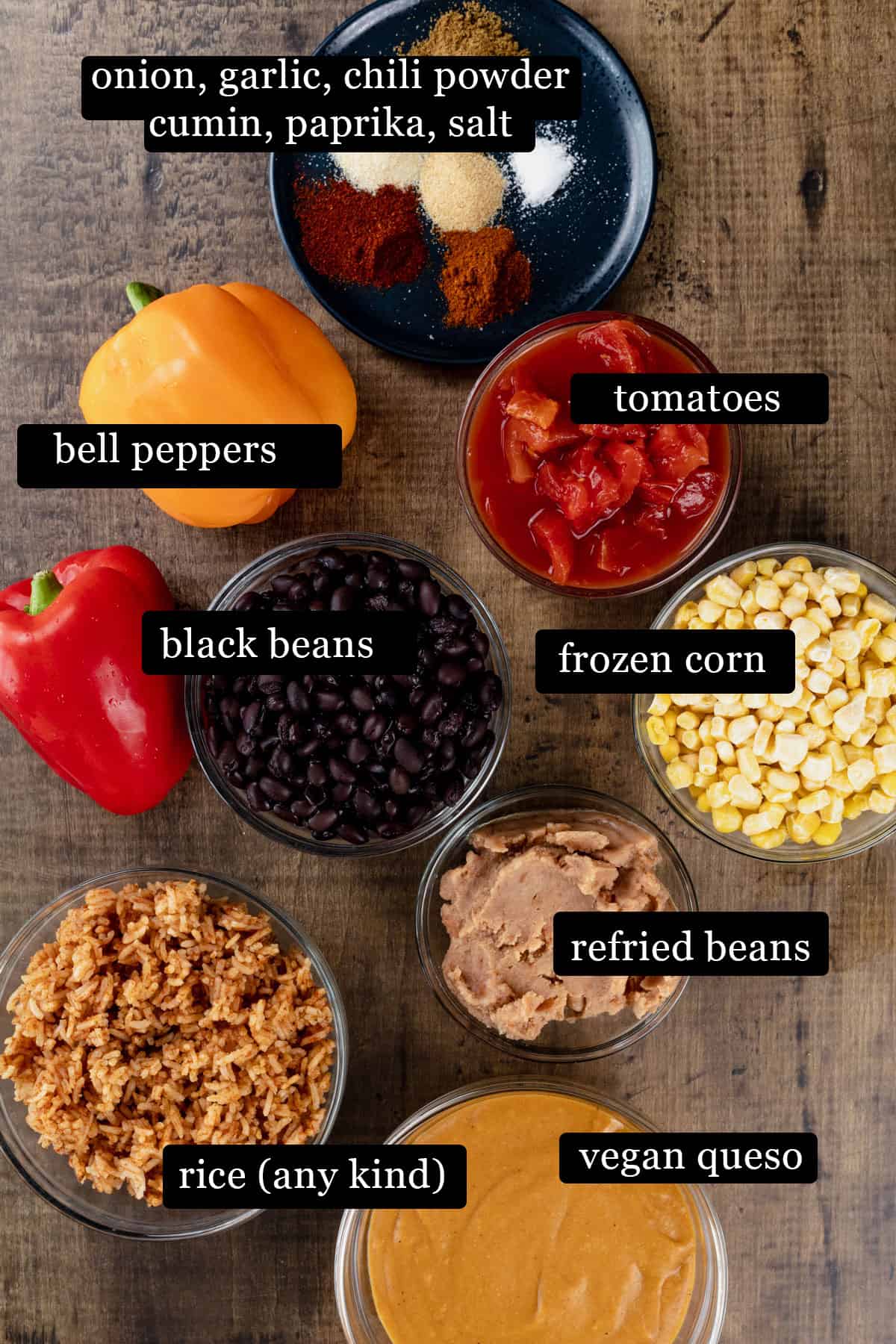 Ingredients for the taco casserole in various glass bowls on a wood tabletop. Black and white labels have been added to name each ingredient like "black beans" and "frozen corn".