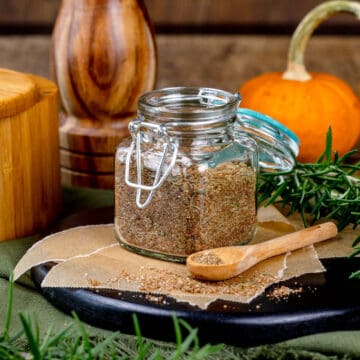 Closeup of a small glass spice jar filled with a turkey seasoning mix. A small wood spoon, fresh rosemary, and mini pumpkins are next to and behind the jar.